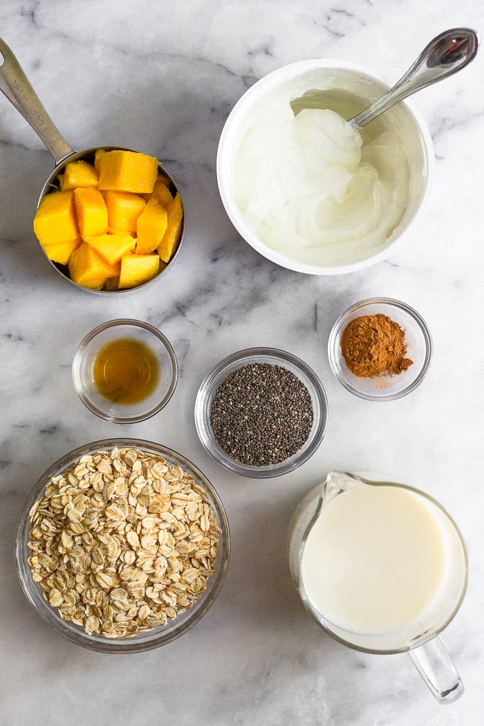 White counter with a container of greek yogurt, glass of almond milk, bowl of rolled oats, cup of diced mangos, bowl of chia seeds, bowl of cinnamon, and a bowl of vanilla extract.