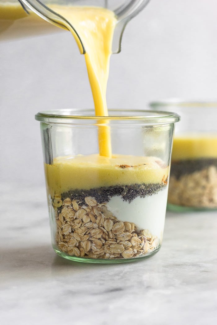 Glass jar filled with rolled oats, greek yogurt, and chia seeds with a blended mango milk being poured into it.