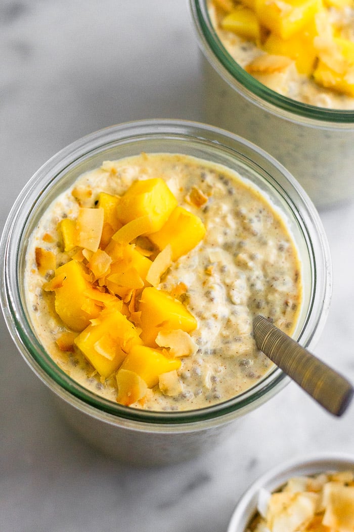 Overnight Oats with Greek yogurt topped with diced mango and shredded coconut. A spoon is coming out of the jar and next to it is a small cup of more coconut and behind it another jar of healthy overnight oats.