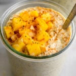 Glass jar filled with mango overnight oats toped with fresh mangos and coconut. There is a spoon in the jar with another jar behind it.
