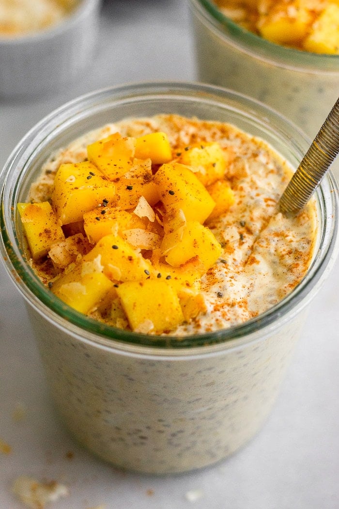 Glass jar filled with mango overnight oats toped with fresh mangos and coconut. There is a spoon in the jar with another jar behind it.