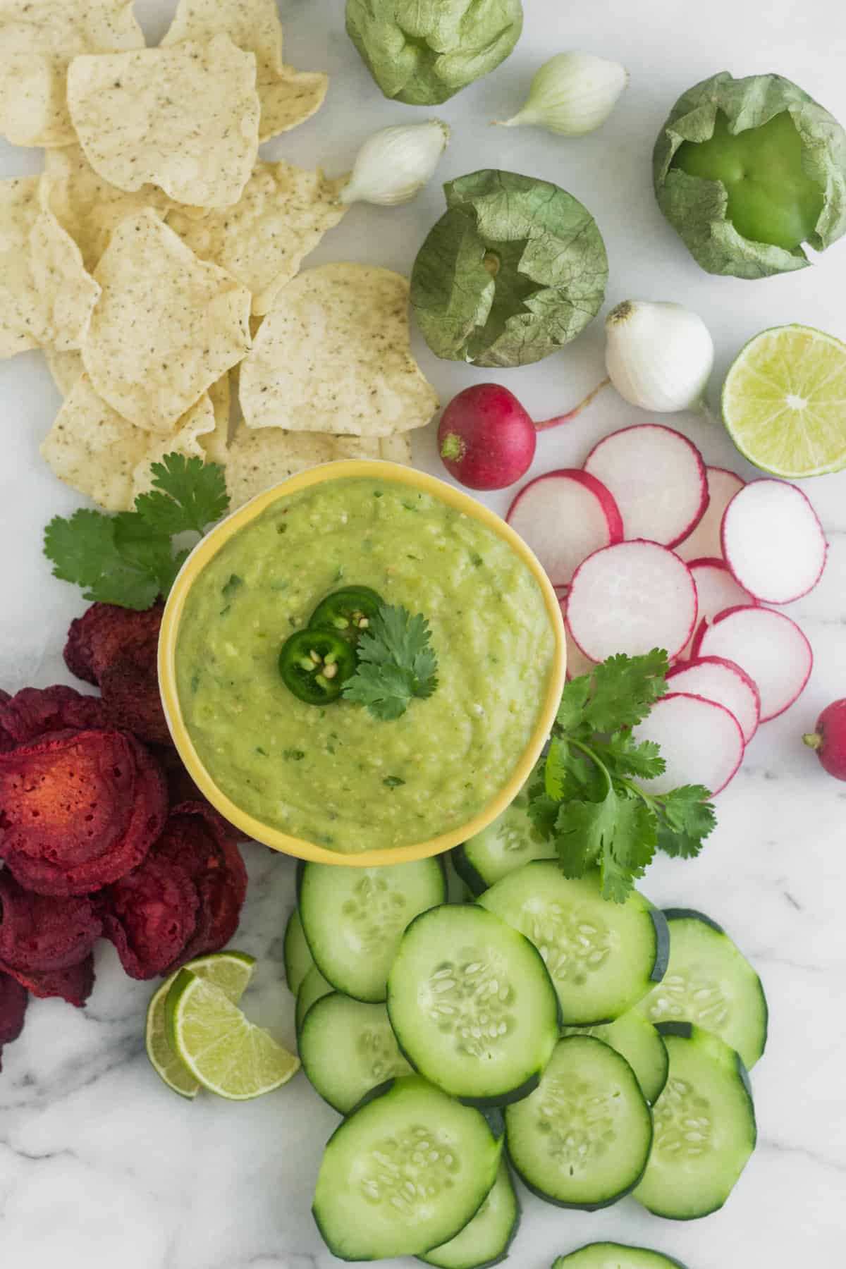 Yellow bowl with Roasted Tomatillo Avocado Salsa surrounded by sliced cucumbers, radishes, beets, chips, and tomatillos