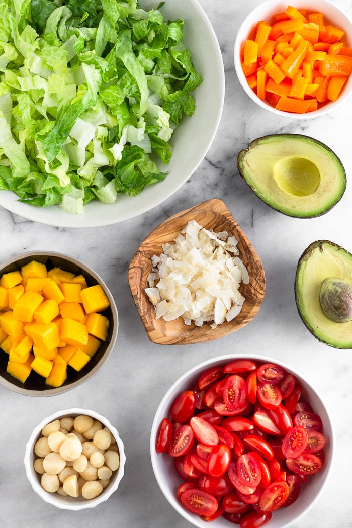 White counter with a big bowl of chopped lettuce, bowl of peppers, an avocado cut open, bowl of halved tomatoes, bowl of macadamia nuts, bowl of diced mango, and a plate of coconut flakes.
