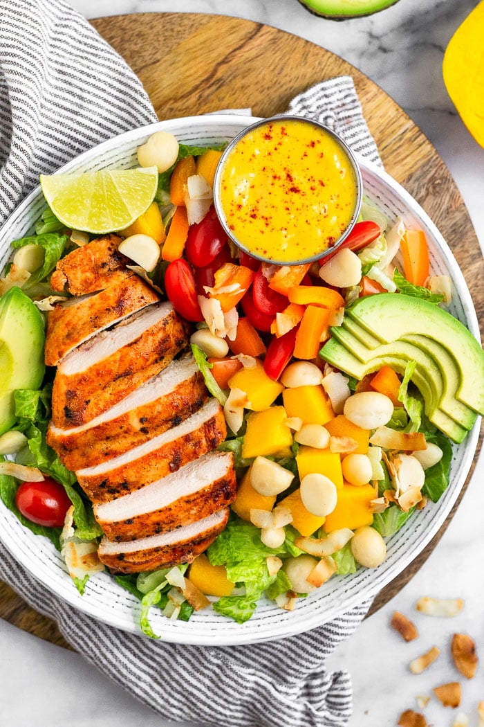 Overheat shot of a tropical chicken salad in large bowl sitting on a towel on a cutting board. It is surrounded by toasted coconut flakes, half a mango, and half an avocado.