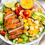 Large bowl of tropical chicken salad with grilled chicken, mango, macadamia nuts, avocado, tomatoes, peppers, and coconut flakes on a bed of lettuce. It is sitting on stripped towel on a circular cutting board with a mango behind it.