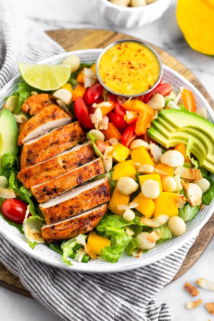 Large bowl of tropical chicken salad with grilled chicken, mango, macadamia nuts, avocado, tomatoes, peppers, and coconut flakes on a bed of lettuce. It is sitting on stripped towel on a circular cutting board with a mango behind it.