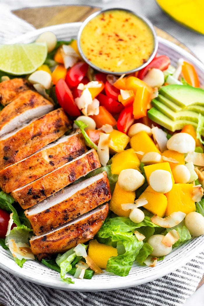 Close up of a large bowl of tropical chicken salad with grilled chicken, mangos, coconut, avocado, tomatoes, peppers, and shredded coconut. There is a small ramekin of coconut mango dressing in the bowl. A mango is behind the bowl.
