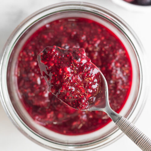 Overhead shot of a jar with berry chia jam with a spoonful of it overtop. Next to is a small bowl of chia seeds and a bowl of frozen berries.