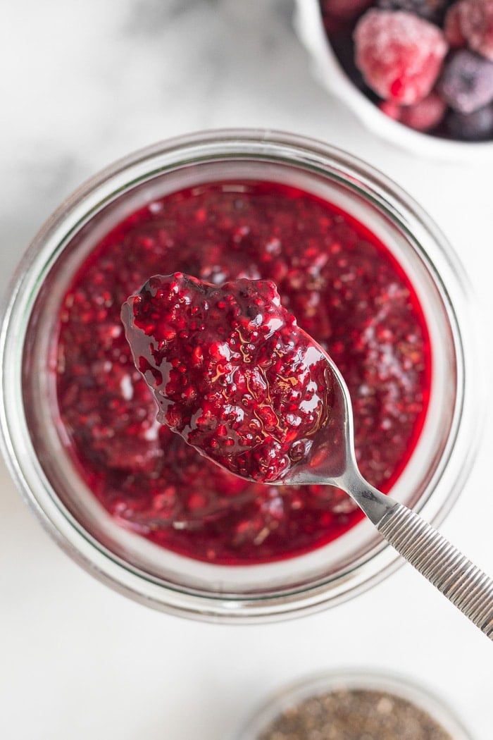 Overhead shot of a jar with berry chia jam with a spoonful of it overtop. Next to is a small bowl of chia seeds and a bowl of frozen berries.