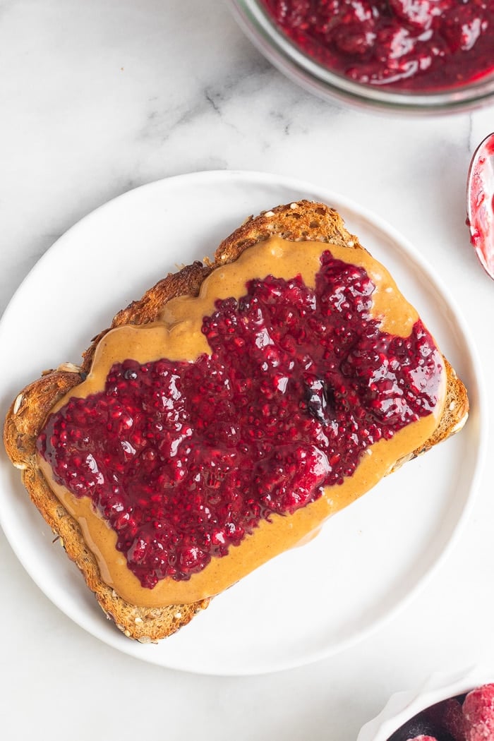 A plate with a piece of toast with peanut butter and mixed berry jam on it. Off in the corner is a large jar with more jam.