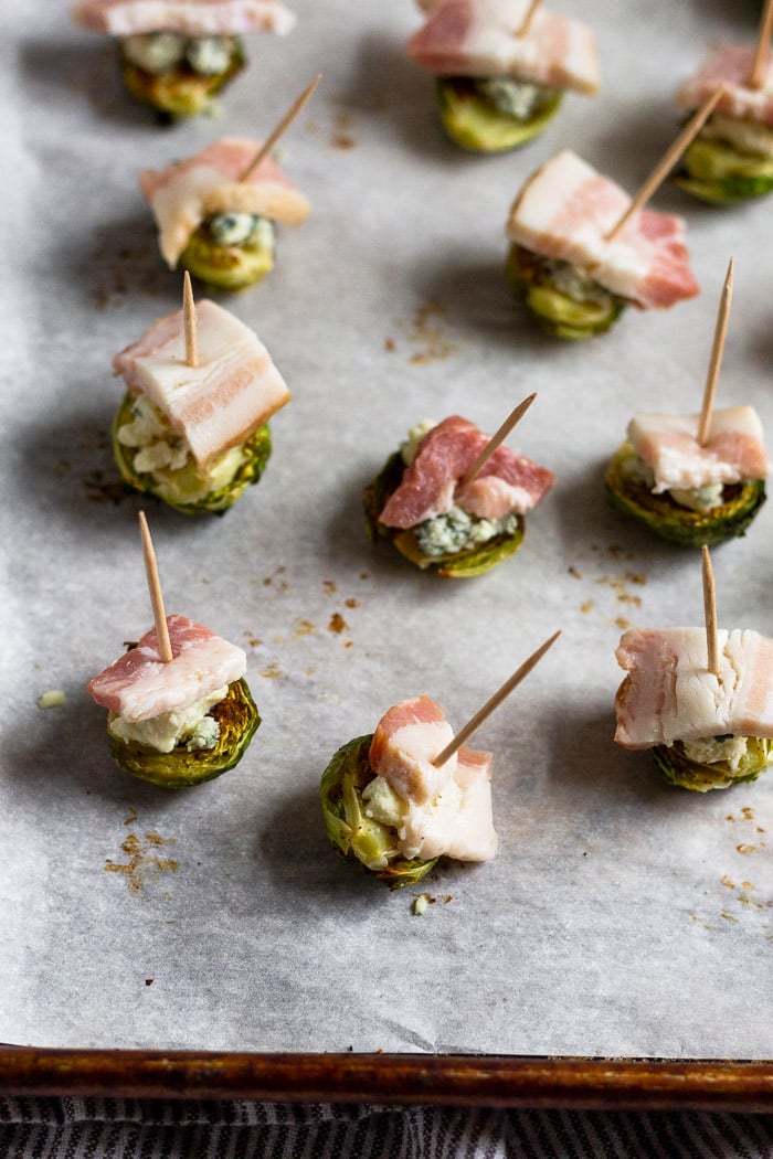 Individual roasted Brussel sprouts topped with blue cheese and a piece of raw bacon on a baking sheet. A toothpick is stuck in each of them.