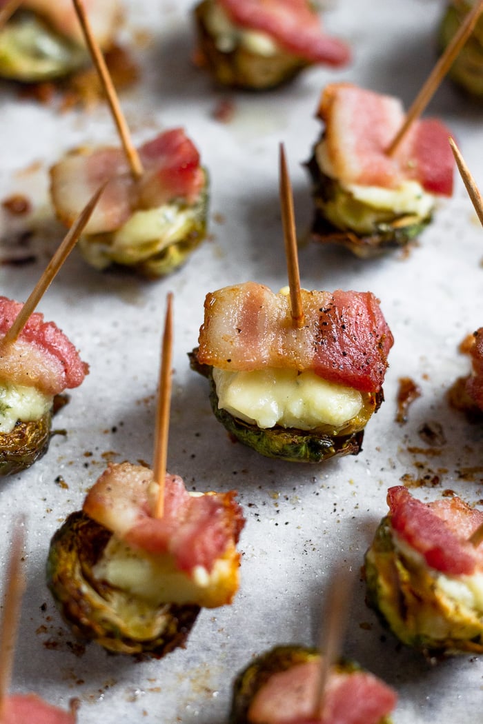 A bunch of crispy roasted brussel sprouts topped with blue cheese and bacon scattered on a baking sheet. A toothpick is stuck in each one.