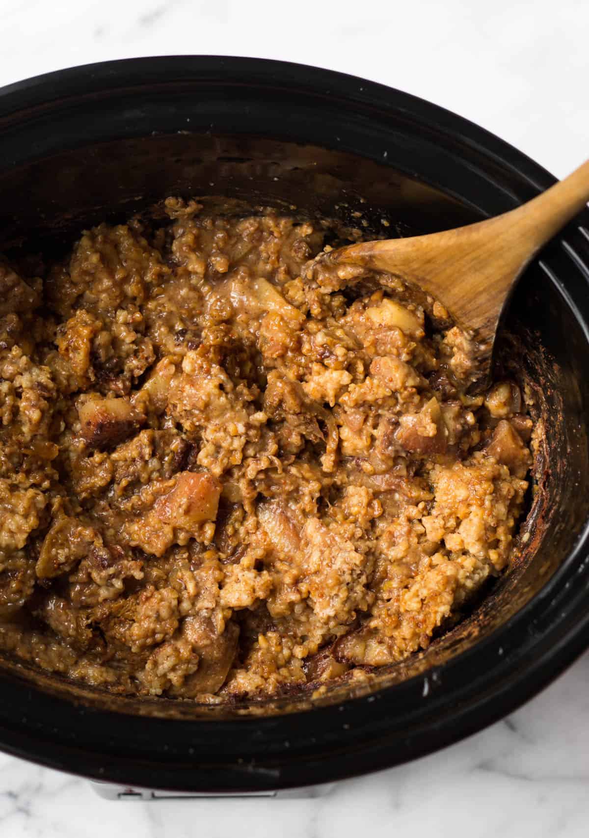 Slow cooker filled with cooked fig apple cinnamon steel cut oats with a wooden spoon it in.