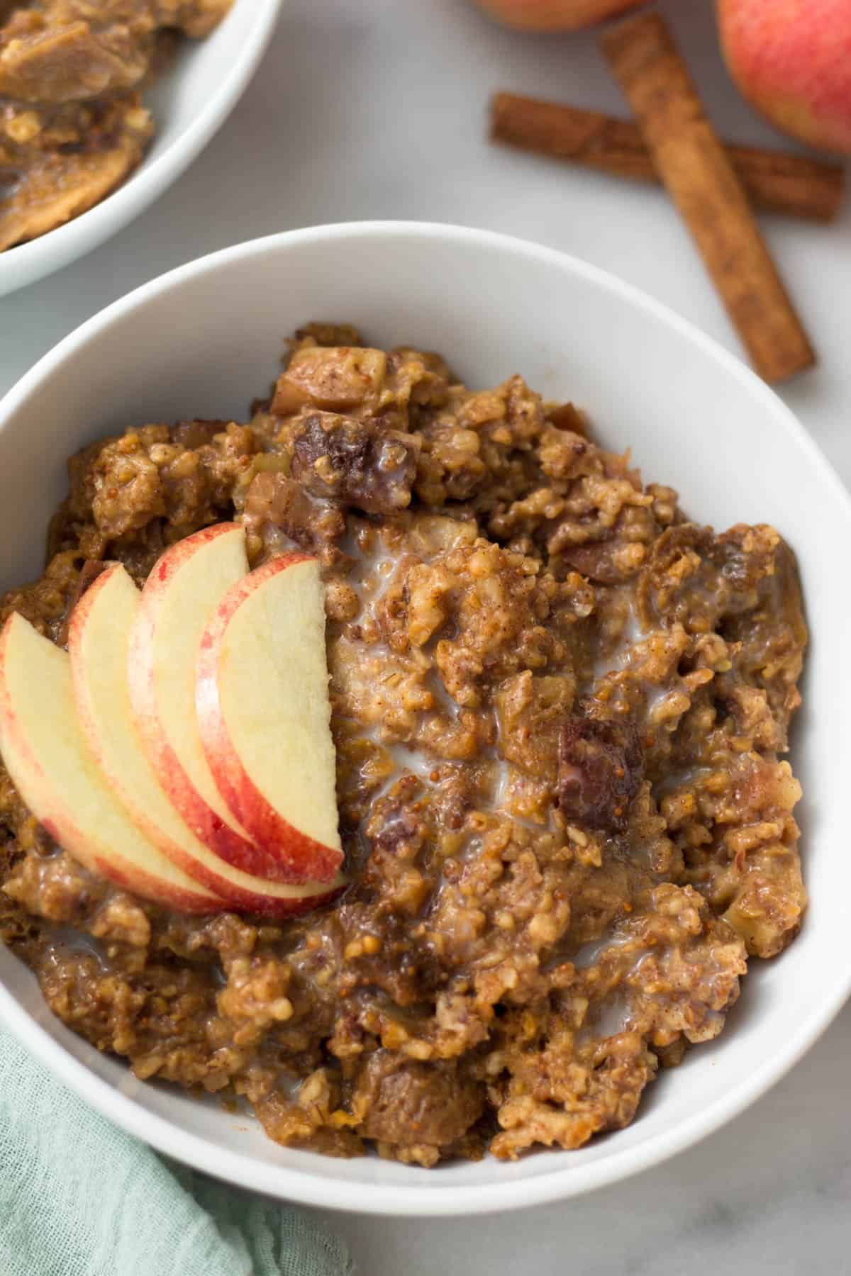 White bowl with vegan Fig Apple Cinnamon Slow Cooker Steel Cut Oats topped with fresh sliced apples. Behind it is cinnamon sticks.