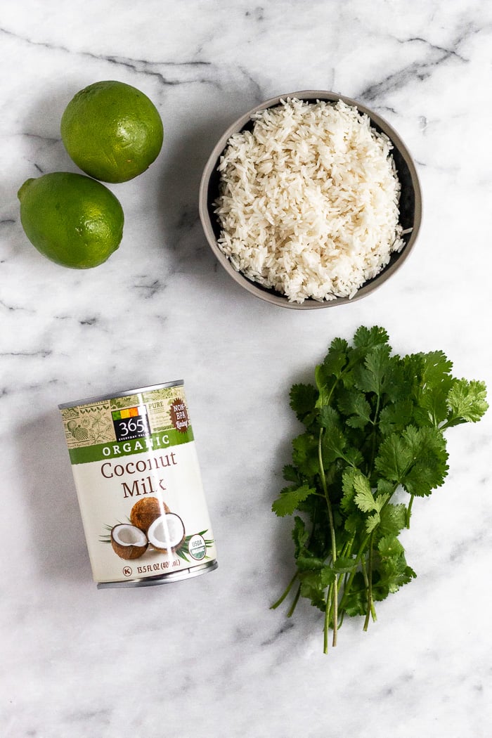 A can of full fat coconut milk. 2 limes, bowl of uncooked white rice, and a small bunch of cilantro on a white counter top to make coconut cilantro lime rice.