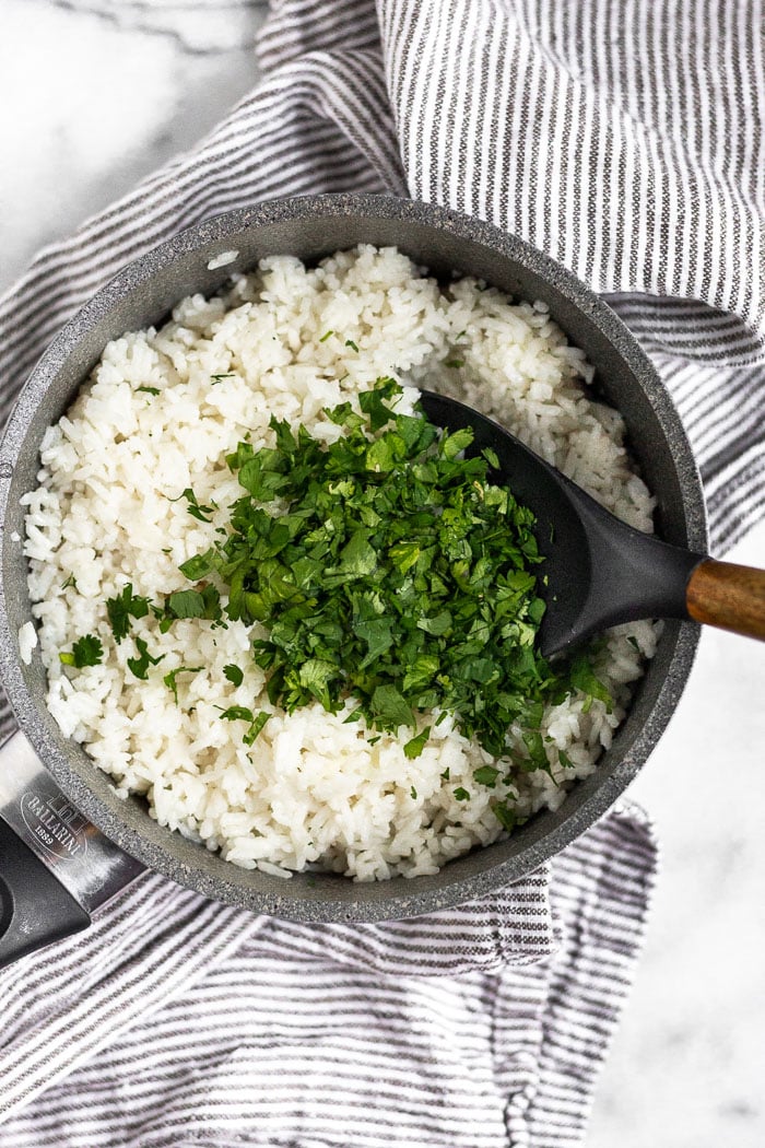Sauce pan filled with cook coconut rice with a large pile of chopped cilantro on top of it. A spoon is sticking out of the pot.
