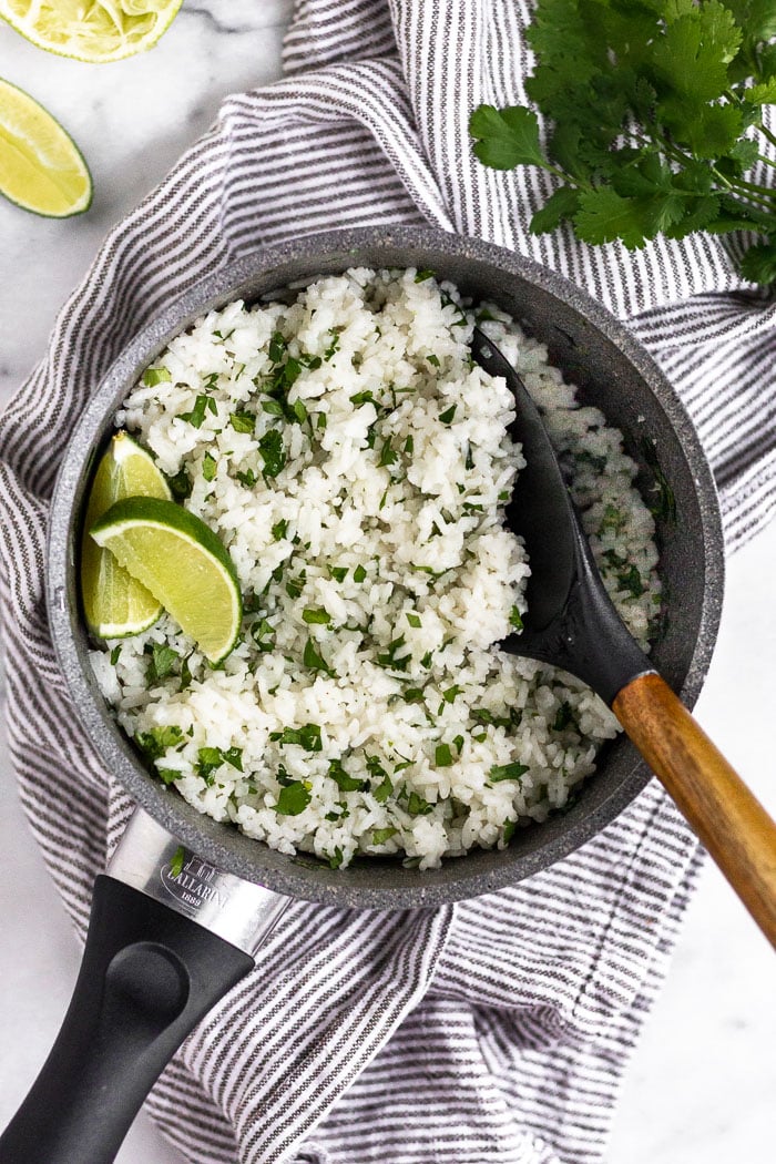Small sauce pan filled with coconut cilantro lime rice garnished with lime wedges. Around it is more limes and cilantro.