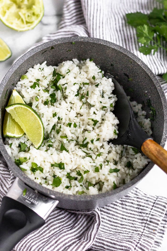 A small pot filled with cooked coconut cilantro lime rice garnished with lime wedges. The pot is sitting on a stripped towel and around it is more limes and cilantro.