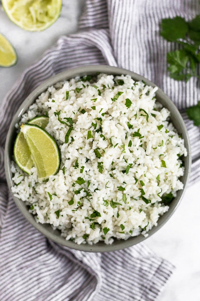 A bowl filled with coconut cilantro lime rice garnished with two lime wedges and surrounded by more limes and cilantro.
