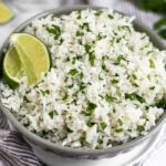 Coconut cilantro lime rice in a grey bowl garnished with lime wedge. The bowl is sitting on a small white plate on a towel with limes and cilantro behind it.