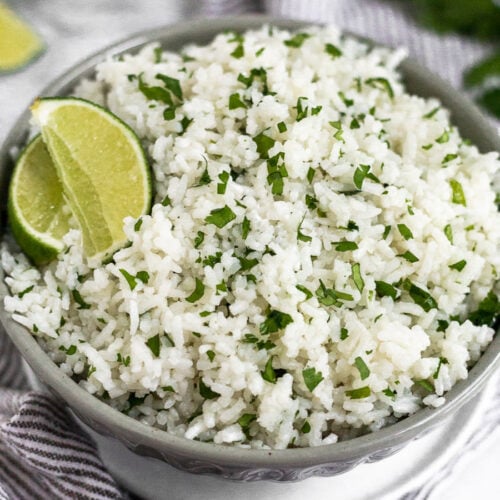 Coconut cilantro lime rice in a grey bowl garnished with lime wedge. The bowl is sitting on a small white plate on a towel with limes and cilantro behind it.