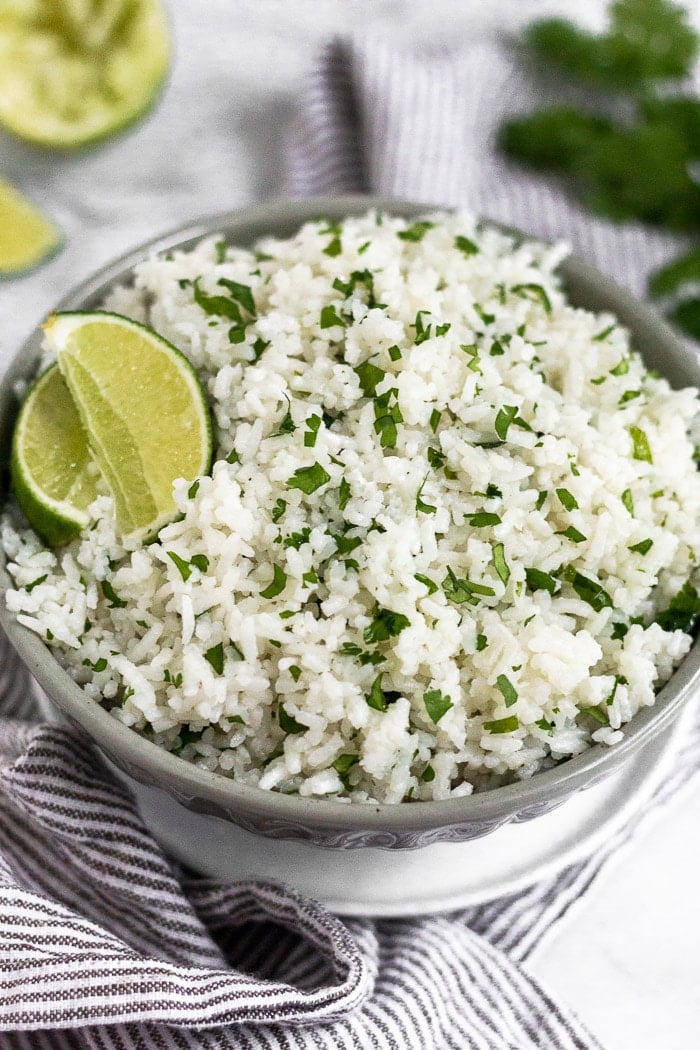 Coconut cilantro lime rice in a grey bowl garnished with lime wedge. The bowl is sitting on a small white plate on a towel with limes and cilantro behind it.