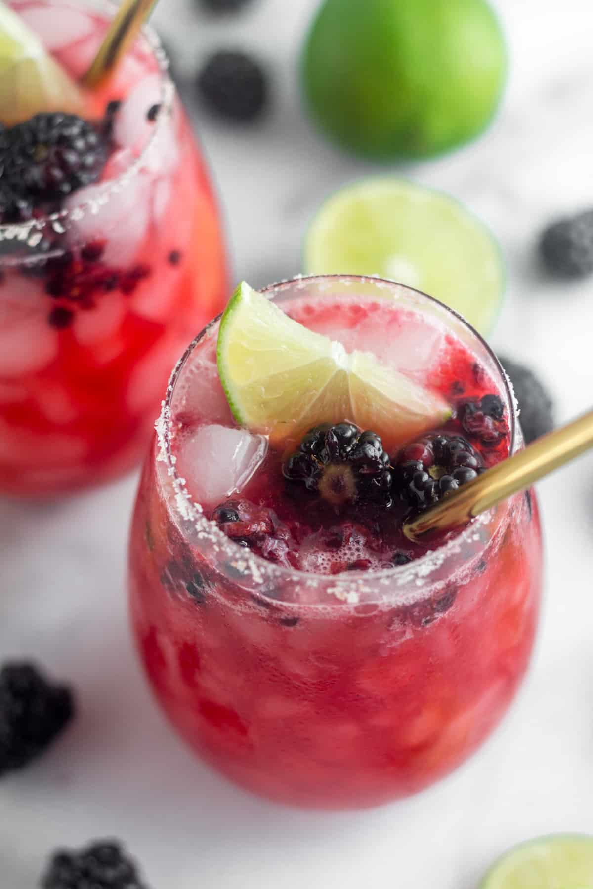 Overhead of a Blackberry Margarita with a lime wedge, blackberries, and a gold straw in it. It is surrounded by blackberries, lime wedges, and another filled glass.