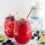 Two glasses filled with blackberry margaritas topped with blackberries and lime wedges. A straw is coming out of both. Around them is more blackberries and lime wedges and a bottle of tequila is behind them.
