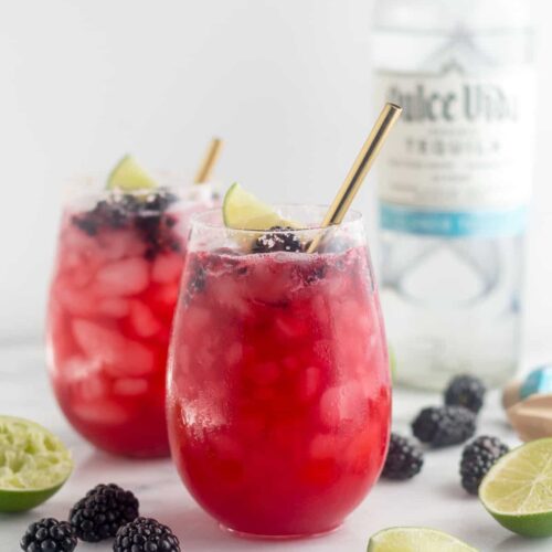 Two glasses filled with blackberry margaritas topped with blackberries and lime wedges. A straw is coming out of both. Around them is more blackberries and lime wedges and a bottle of tequila is behind them.