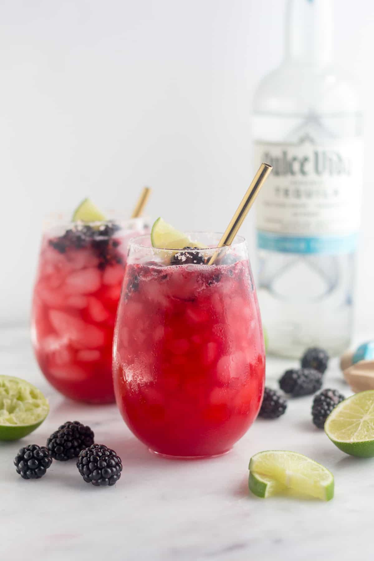 Two glasses filled with blackberry margaritas topped with blackberries and lime wedges. A straw is coming out of both. Around them is more blackberries and lime wedges and a bottle of tequila is behind them.