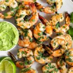 White plate filled with easy grilled shrimp skewers with avocado sauce and lime wedges.