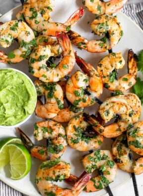 White plate filled with easy grilled shrimp skewers with avocado sauce and lime wedges.