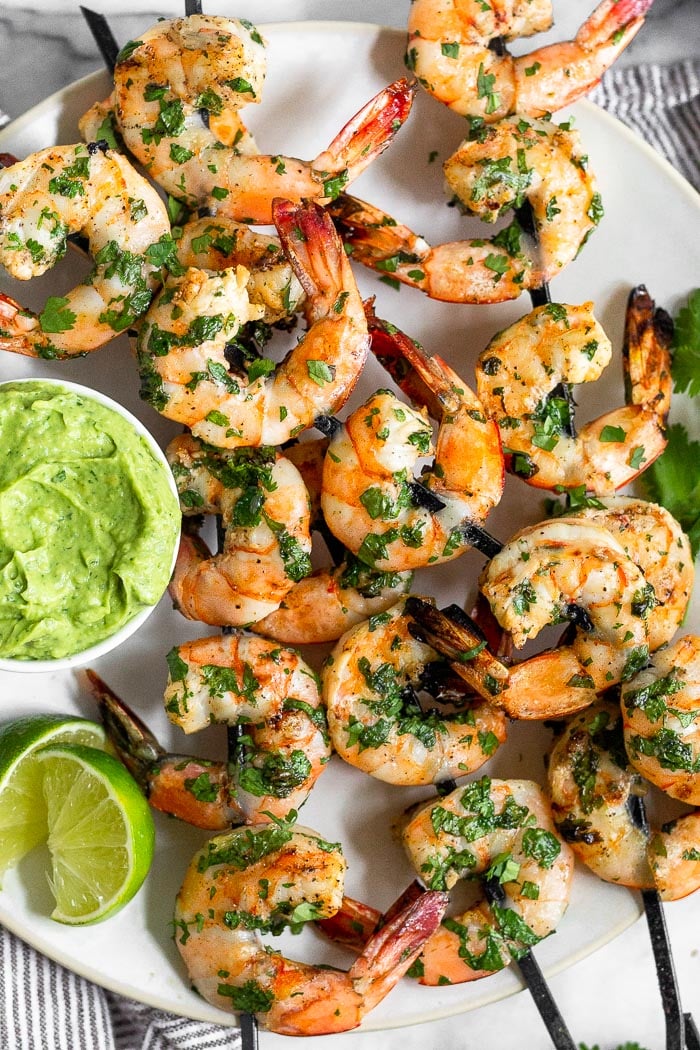 White plate filled with easy grilled shrimp skewers with avocado sauce and lime wedges.