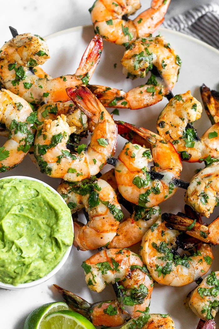 Plate of grilled shrimp skewers with a dish of avocado sauce and lime wedges on it.