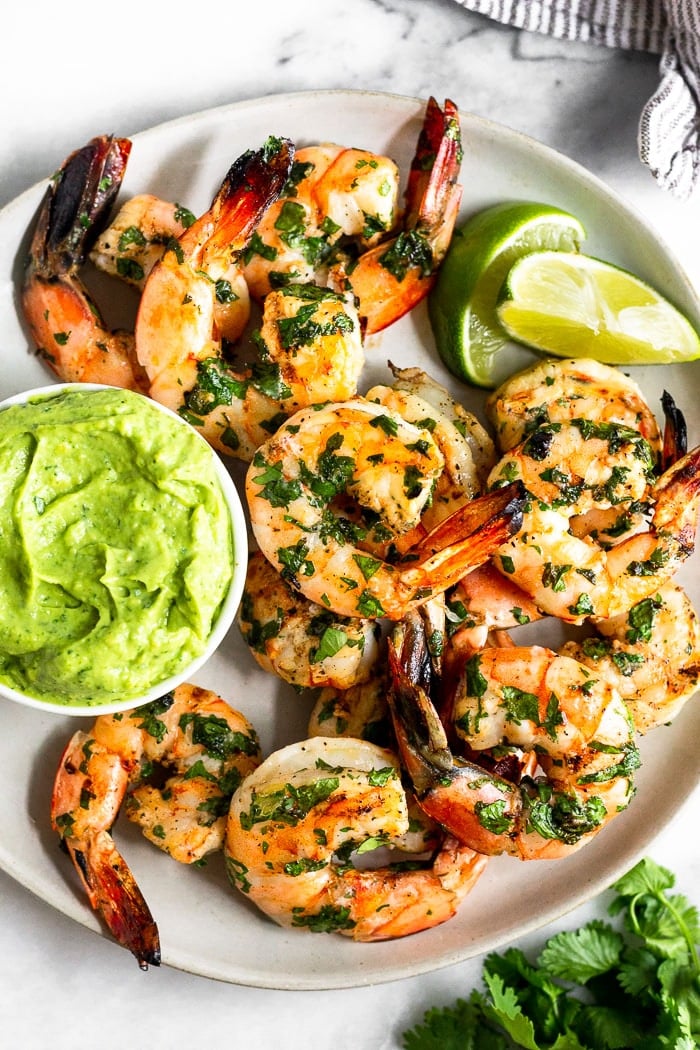 Plate of grilled cilantro lime shrimp with a small dish of avocado sauce and lime wedges. A bunch of cilantro and a kitchen towel is around it.