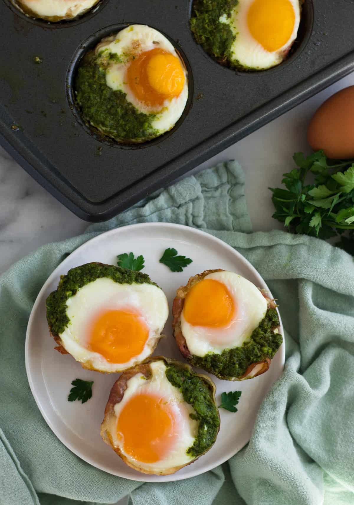 Overhead of 3 prosciutto egg cups on a white plate and a muffin tin with more egg cups