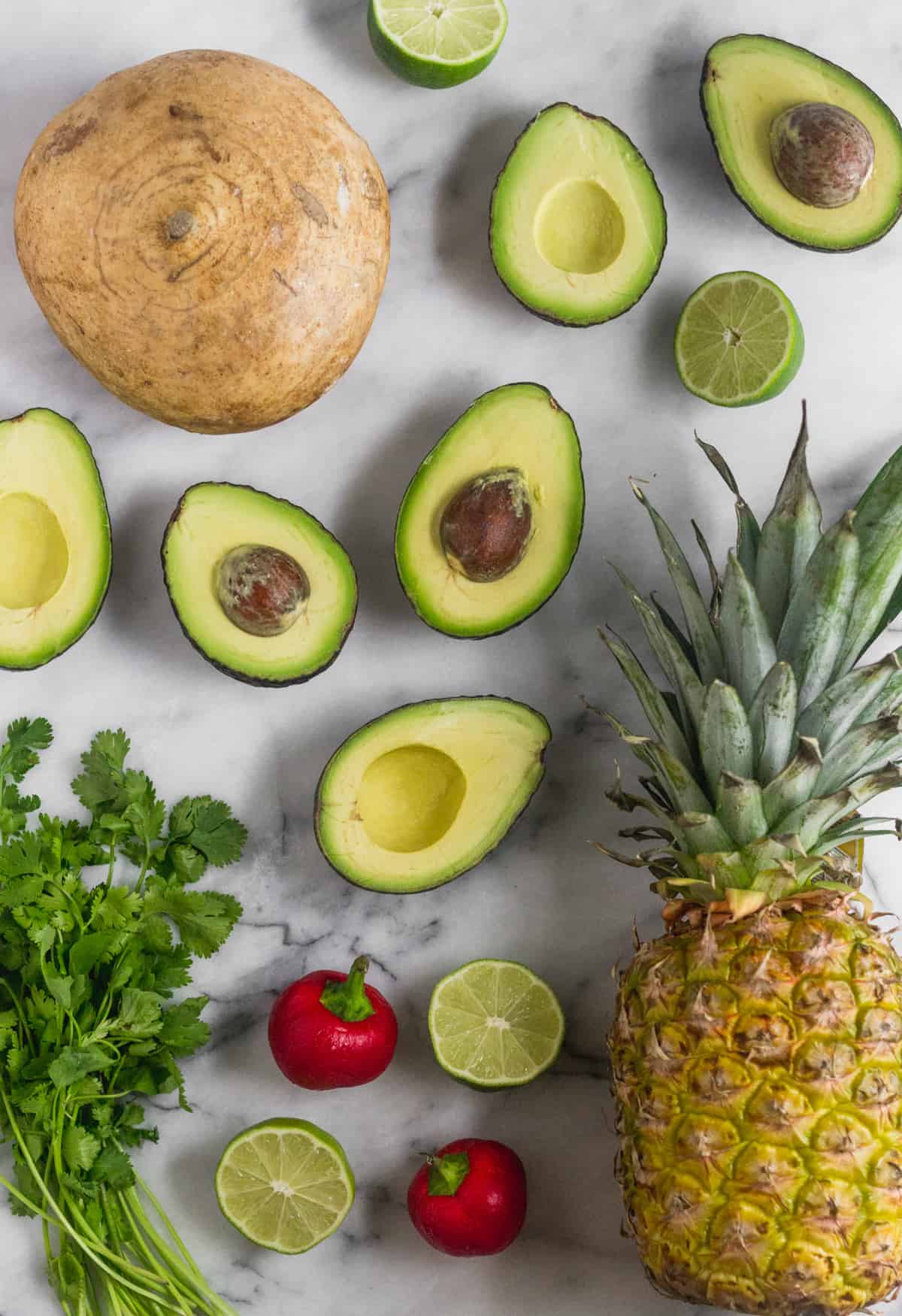 White counter with a jicama, halved avocados, halved limes, a while pineapple, cherry peppers, and cilantro on it