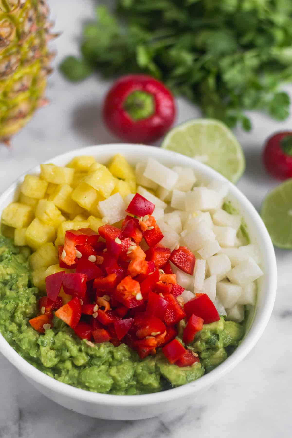 White bowl filled with mashed avocado, diced pineapple, diced jicama, and diced cherry peppers, with limes, cherry peppers, cilantro, and a pineapple behind it