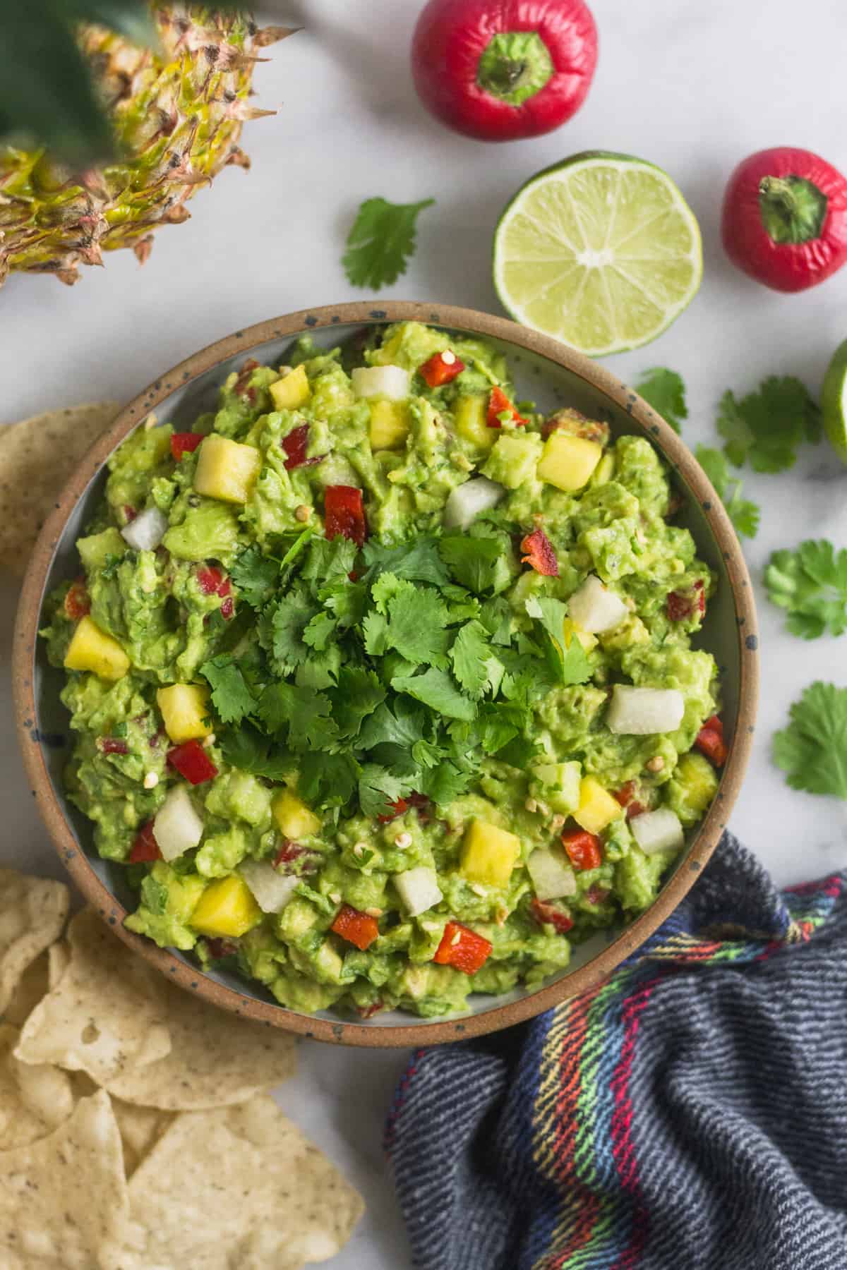 Bowl of pineapple jicama guacamole surrounded by chips, whole pineapple, limes, cherry peppers, cilantro leaves, and a festive towel