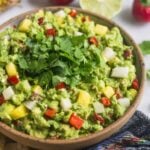 Large bowl filled with pineapple jicama guacamole with a blue towel, chips, limes, and cherry peppers around it