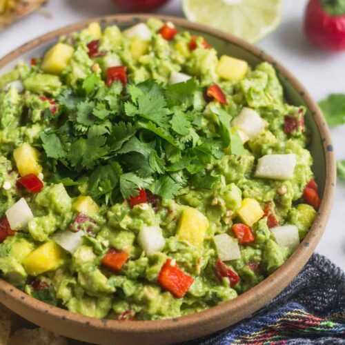 Large bowl filled with pineapple jicama guacamole with a blue towel, chips, limes, and cherry peppers around it