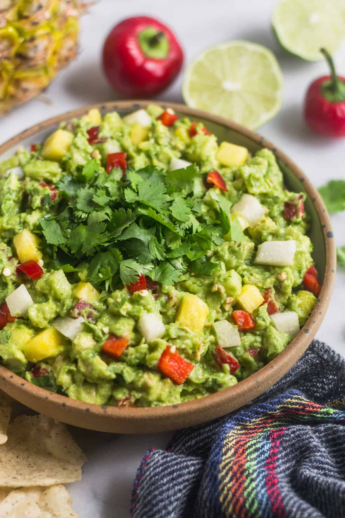 Large bowl filled with pineapple jicama guacamole with a blue towel, chips, limes, and cherry peppers around it