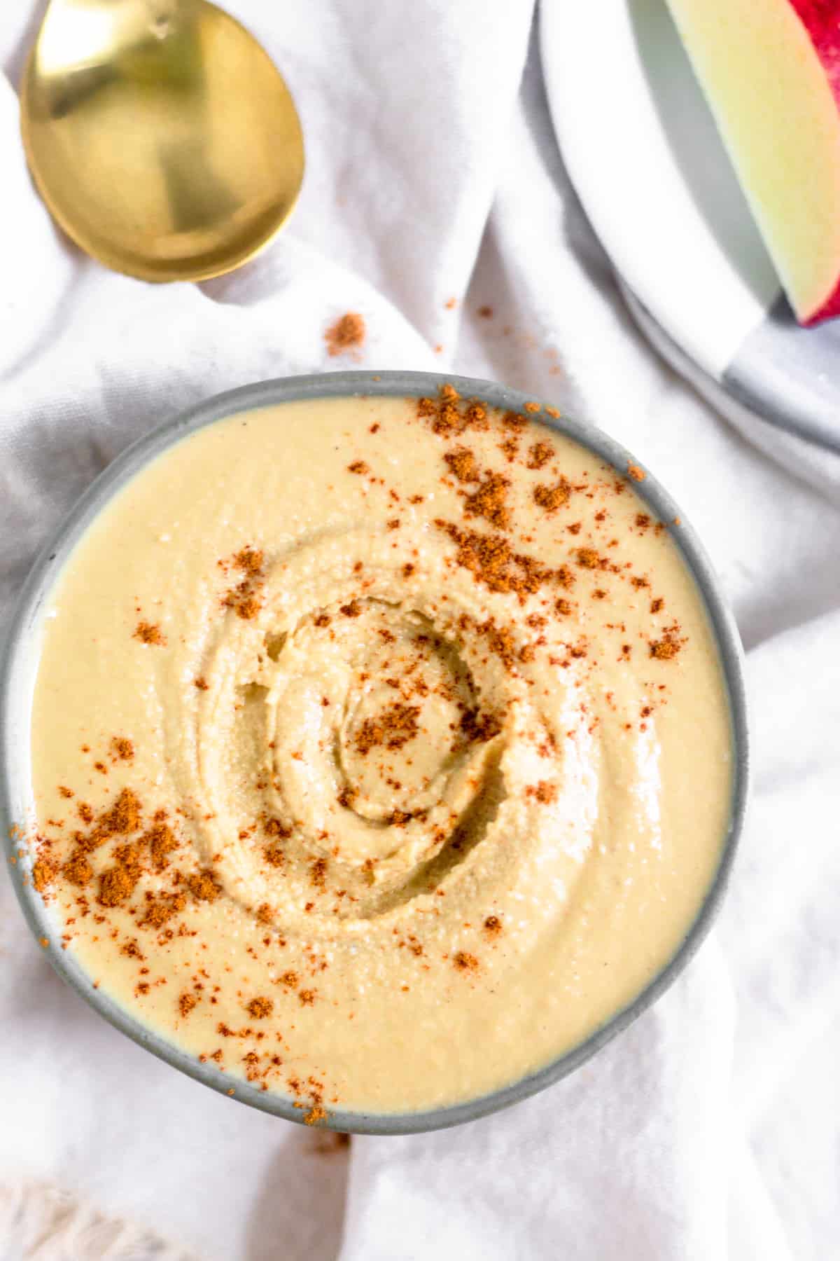 Overhead of a small bowl on a white linen filled with cashew coconut butter sprinkled with cinnamon. Next to it is a aptly of sliced red apples and a gold spoon.