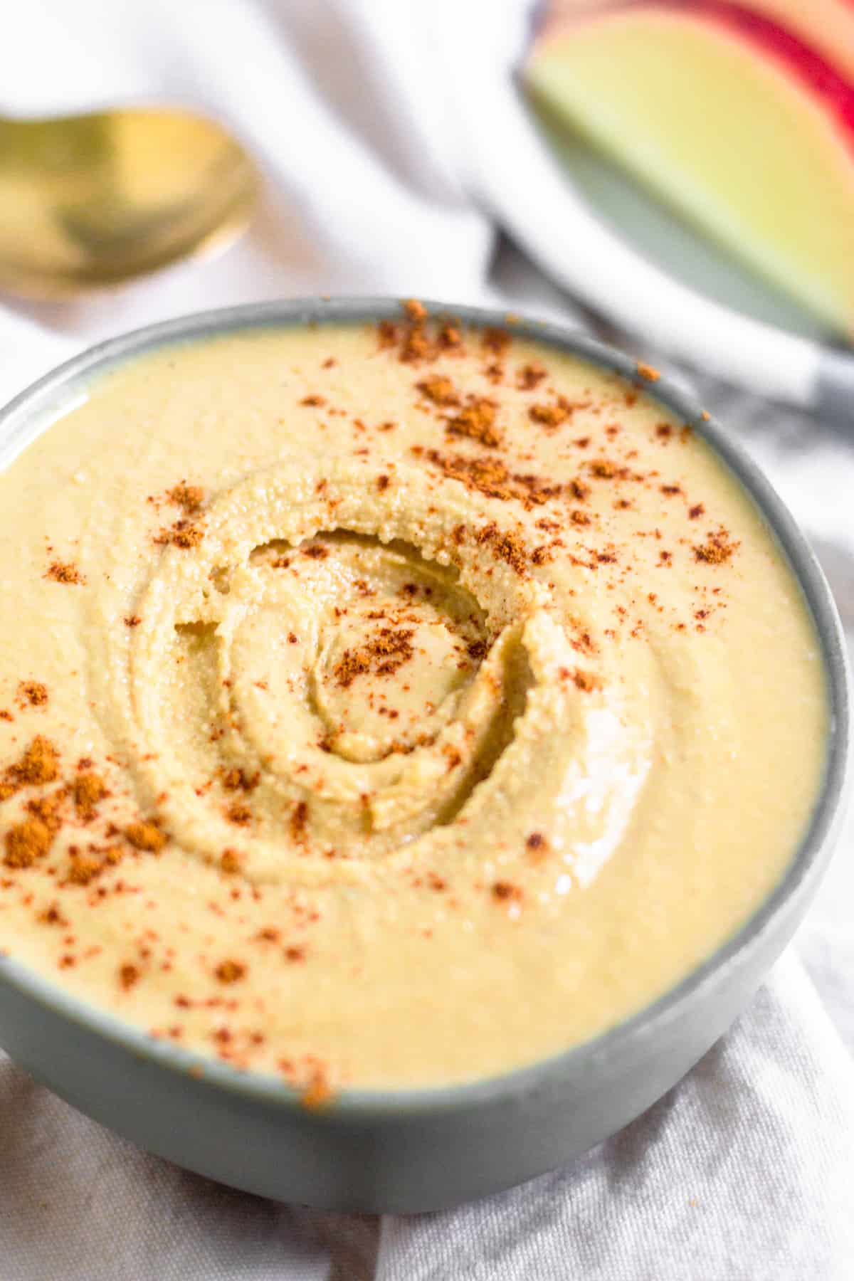 Homemade cashew coconut butter in a green small bowl sprinkled with cinnamon. Behind it is a plate of sliced apples and a spoon