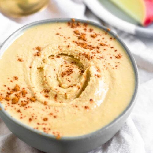 Cashew coconut butter in a green small bowl on a white linen sprinkled with cinnamon. Behind it is a plate of sliced apples and a spoon