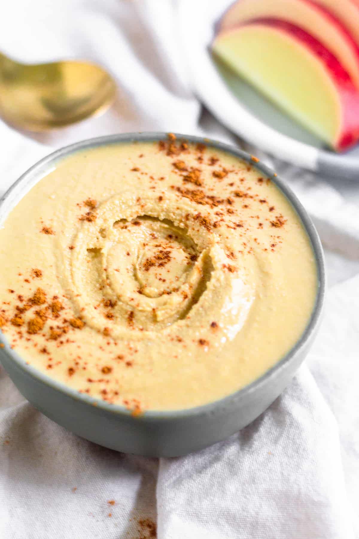 Cashew coconut butter in a green small bowl on a white linen sprinkled with cinnamon. Behind it is a plate of sliced apples and a spoon