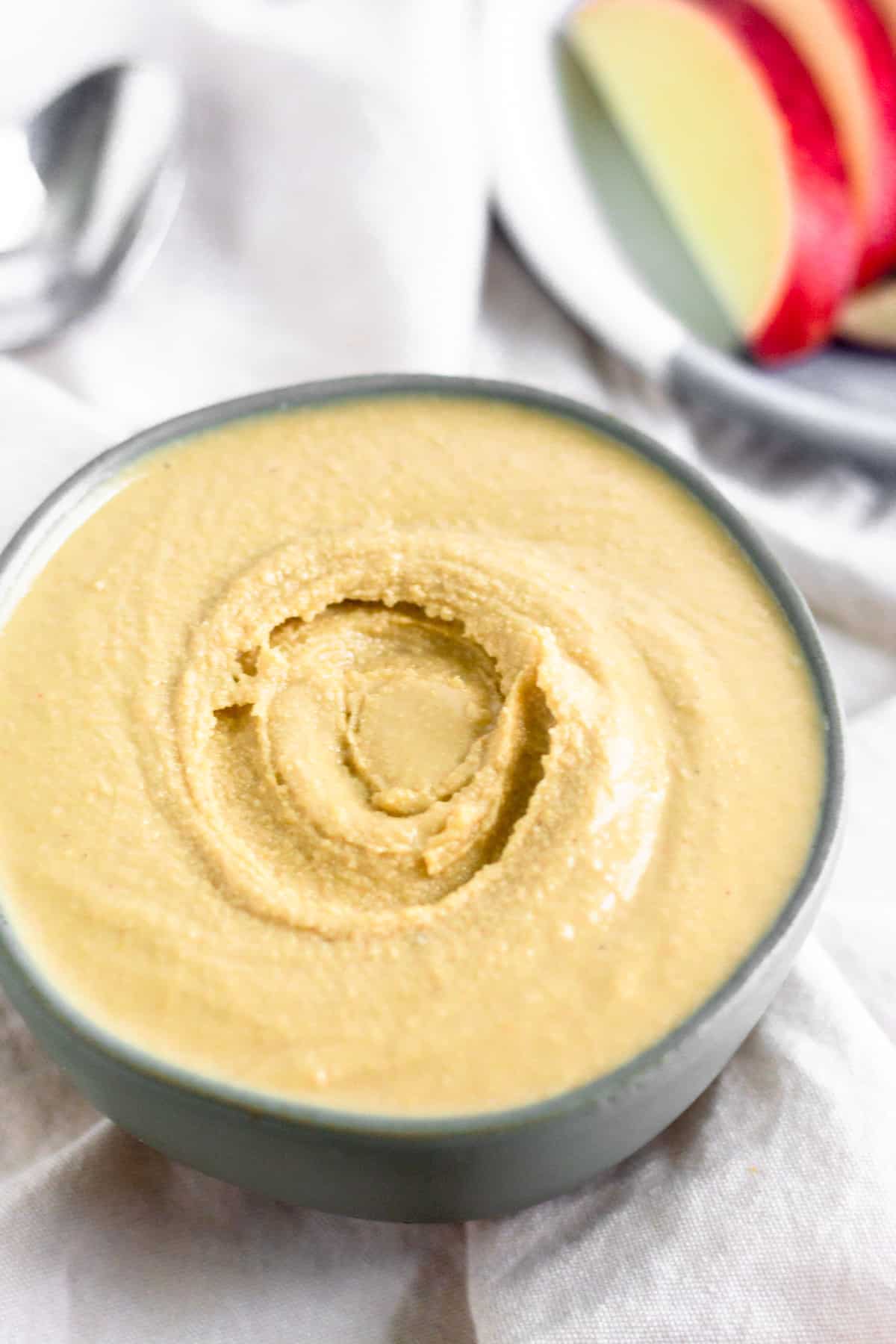 A bowl of freshly made cashew coconut butter on a white linen with a plate of sliced apples and a spoon behind it