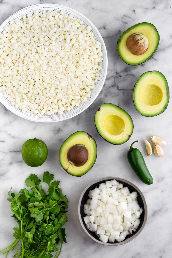 White counter with a bowl of cauliflower rice, sliced avocados, jalapeno, garlic, a bowl of diced onion, a bunch of cilantro, and a lime.
