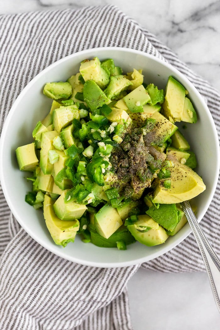 Bowl of diced avocado, chopped jalapenos, lime juice, and salt and pepper. There is spoon sticking out of the bowl and it is sitting on a stripped towel.