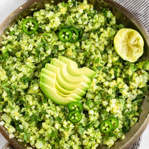 A large pan filled with avocado cauliflower rice topped with sliced jalapeños, avocado slices, and half a lime. The pan is sitting on a towel and around it is half and avocado, cilantro, and sliced japaleons.